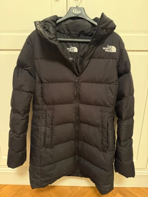 Svart lång pufferjacka The North Face - Säljer en svart lång pufferjacka från The North Face med huva och dragkedja framtill. Jackan har två sidofickor med dragkedja och broderad logga på bröstet och ryggen. Perfekt för kalla dagar.
