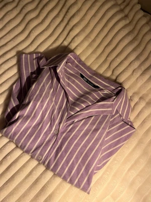 Ljuslila randig skjorta från Zara - Säljer en ljuslila skjorta från Zara med vita vertikala ränder. Skjortan har klassisk krage, långa ärmar med knappar. Perfekt för dig som gillar stilrena och trendiga plagg med lite färg. 