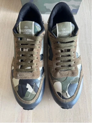 Valentino Garavani Rockrunner sneakers - Säljer ett par Valentino Garavani Rockrunner sneakers med camouflagemönster i grönt, beige, svart och brunt. Skorna har detaljer i mocka och skinn, platt sula och rund tå. Klassisk snörning och röd detalj på hälen. Levereras med originalkartong och extra skosnören.