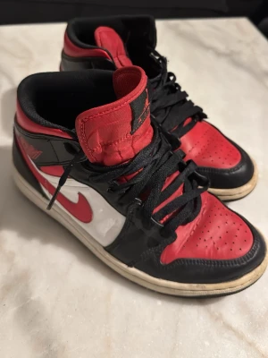 Nike Air Jordan 1 High röd/svart - Säljer ett par Nike Air Jordan 1 High med klassisk färgblockning i rött, svart och vitt. Skorna har snörning, rund tå och platt sula. Materialet är mestadels skinn med textil på plösen. Snygg retrostil och ikonisk design med Jumpman-logga på tungan.