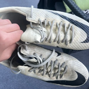 Nike Mercurial vita fotbollsskor - Säljer ett par Nike Mercurial fotbollsskor i vitt med stora svarta swoosh-loggor. Skorna har snörning, rund tå och dobbar för bra grepp på planen. Tillverkade i syntetmaterial och designade för fart och kontroll på fotbollsplanen.
