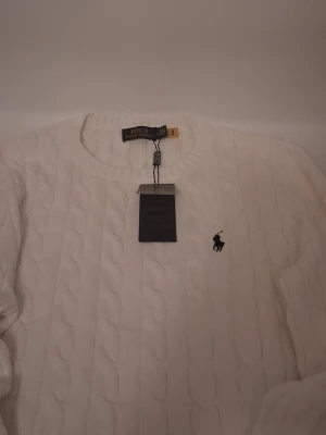 Vit stickad tröja från Polo Ralph Lauren - Vit kabelstickad tröja från Polo Ralph Lauren i storlek M. Tröjan har rund hals, klassisk svart broderad logga på bröstet och ribbade muddar. Perfekt för dig som gillar stilrena och tidlösa plagg med premiumkänsla.