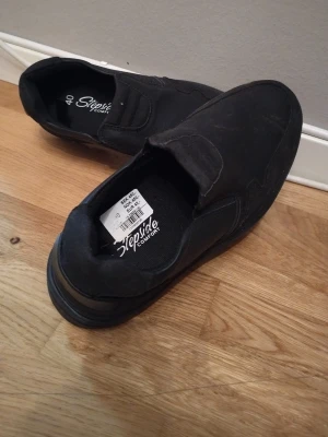 Svarta loafers från Stepside Comfort - Säljer ett par svarta loafers från Stepside Comfort i storlek 40. Skorna har en rund tå och platt sula med mönstrad undersida för extra grepp. De är tillverkade i syntetmaterial och har en enkel slip-on design utan snörning.