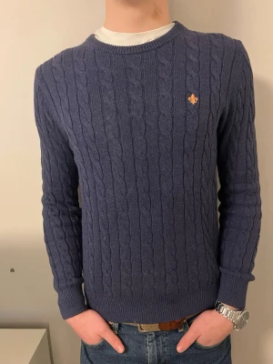 Morris marinblå kabelstickad tröja - Snygg marinblå kabelstickad tröja från Morris med rund hals och diskret logga broderad på bröstet. Tröjan är gjord i mjuk kashmir och har ribbade muddar vid ärmslut och nederkant. Perfekt för dig som gillar klassisk stil med en modern touch.