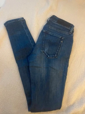Replay New Luz blå jeans, storlek 24/30 - Snygga mörkblå jeans från Replay, modell New Luz. Klassisk femficksdesign med knapp och dragkedja fram. Jeansen har en slim passform och är tillverkade i mjukt bomullsmaterial med lite stretch för extra komfort. Perfekta för dig som gillar stilrena och tidlösa jeans.