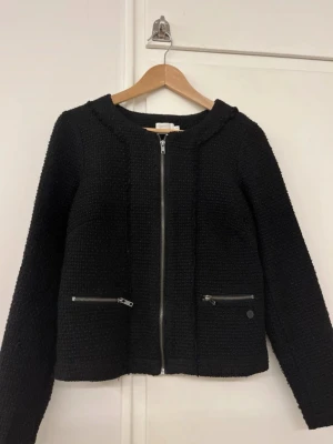 Svart jacka/kofta - Svart jacka/kofta med grovstickad struktur och rund hals. Jackan har två fickor med dragkedjor framtill och en silverfärgad dragkedja som stängning. Långärmad och med en liten metallbricka med PEP-logga vid ena fickan.