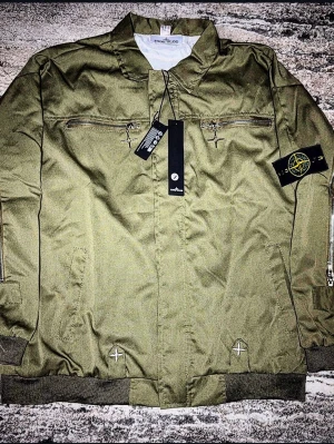Stone Island jacka - Säljer en unik Stone Island-jacka i en snygg olivgrön färg. Jackan har en teknisk design med karakteristiska dragkedjor längs ärmarna och ryggen samt dekorativa broderier. Detaljer: • Skick: Nyskick/Oanvänd med tags kvar. • Funktioner: Rejäla dragkedjor, klassisk Stone Island-patch på ärmen och muddar vid midja/ärmslut. • Passform: Storlek L En riktigt cool samlaritem eller för dig som vill ha en Stone Island-jacka som sticker ut från mängden. Skickas spårbart på plick.