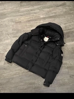 Svart pufferjacka från Moncler - Säljer en svart pufferjacka från Moncler med huva och logotyp på ärmen. Jackan har dragkedja och knappar framtill, samt två sidofickor. Den är vadderad och har en schysst oversized look som passar perfekt när det är kallt ute.