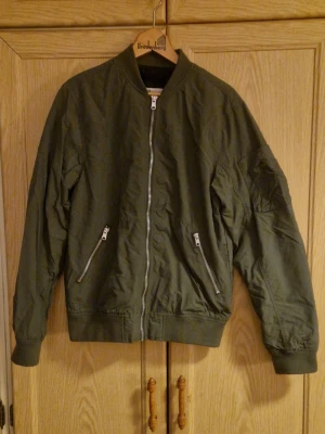 Olivgrön bomberjacka från H&M - Snygg olivgrön bomberjacka från H&M med klassisk ribbad krage och muddar. Jackan har dragkedja framtill, två snedställda fickor med dragkedja och en extra ficka på ärmen. Perfekt för dig som gillar streetstyle och vill ha en chill look.