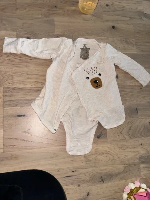 Beige baby body med björntryck från Lindex Lindex vit body, storlek 62-68 - Söt beige baby body från Lindex i storlek 62. Västen har ett gulligt björnansikte tryckt framtill och praktiska tryckknappar längs sidan. Tillverkad i mjuk bomull med lite stretch för extra komfort. Perfekt för små äventyrare!Beige baby body med björntryck från Lindex Lindex vit body, storlek 62-68