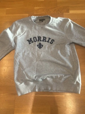 Grå sweatshirt från Morris, storlek M - Säljer en grå sweatshirt från Morris i storlek M. Tröjan har rund hals, ribbade muddar vid ärmslut och nederkant samt ett mörkblått tryck med Morris-logga på bröstet. Materialet är mjuk bomull och polyester, perfekt för chill dagar.