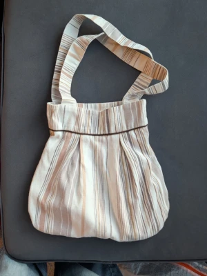 Randig axelväska i bomull - Säljer en mjuk axelväska med randigt mönster i beige och bruna toner. Väskan har breda axelband och är tillverkad i bomullstyg, med rymligt innerfack. Perfekt för att bära med sig allt man behöver under dagen.