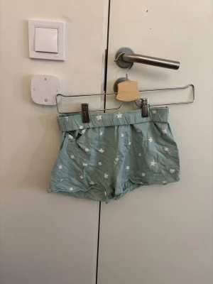 Mintgröna shorts med stjärnmönster - Säljer ett par mintgröna shorts med vita stjärnor. De har en loose passform och elastisk midja för extra komfort. Perfekta för chill dagar och häng hemma. Materialet känns mjukt och lätt, troligen bomull. Snyggt mönster som sticker ut!