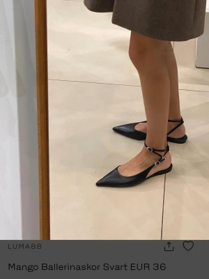 Svarta ballerinaskor från Mango - Säljer ett par svarta ballerinaskor från Mango i storlek 36. Skorna har spetsig tå, smala remmar med spänne runt ankeln och platt sula. Tillverkade i syntetmaterial med stilren och minimalistisk design som ger en modern vibe.