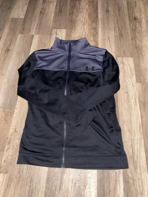 Svart och grå tracksuit Under Armour - Snygg zip-tröja från Under Armour i svart med grått parti över axlar och krage. Tröjan har lång ärm, hög krage och hel dragkedja framtill. Perfekt för träning eller chill, med diskret logga på bröstet. Materialet är mjukt och syntetiskt för skön komfort. (Byxor kommer med tröjan) Ny pris 700