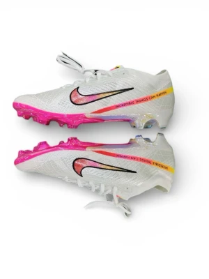 Nike Mercurial vita fotbollsskor - Nike Mercurial fotbollsskor i vit med färgstark rosa och gul sula, svart swoosh och texten 'INCREDIBLE THINGS CAN HAPPEN' på sidan. Skorna har snörning, platt sula med dobbar och är tillverkade i syntetmaterial. Perfekta för dig som vill sticka ut på planen.