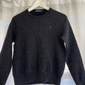 Mörkgrå tröja från Polo Ralph Lauren - Säljer en stilren mörkgrå tröja från Polo Ralph Lauren med lila logga på bröstet. Tröjan är långärmad och har en klassisk rund halsringning. Tröjan är i storlek L men passar en xs. Perfekt för en avslappnad och snygg look. 