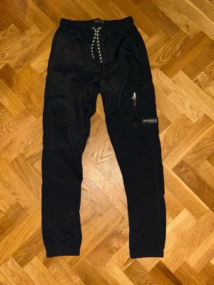 Svarta cargopants från McKenzie - Svarta cargopants från McKenzie med snörning i midjan och dragkedja på benfickan. Byxorna har en loose passform och är gjorda i lätt polyester. Perfekt för dig som gillar streetwear och vill ha praktiska fickor.