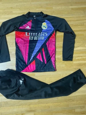 Real Madrid träningsset från Adidas - Säljer ett Real Madrid träningsset från Adidas med långärmad tröja och svarta byxor. Tröjan har coolt grafiskt mönster i rosa, röd, blå och svart med klubbmärke och Adidas-logga. Materialet är syntet, perfekt för träning. Byxorna är helsvarta med Adidas-detalj.