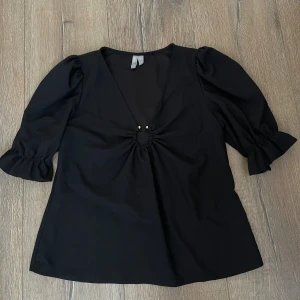 Svart blus med puffärm från ASOS - Säljer en svart blus från ASOS i storlek EU 40. Blusen har puffiga trekvartsärmar med volang vid ärmslut och en djup v-ringning med snygg snörningsdetalj och små pärlor framtill. Materialet känns mjukt och lätt, perfekt för att lyfta din outfit.