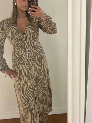 Zebramönstrad långklänning från Adoore - Supersnygg långklänning från Adoore med zebramönster i beige och brunt. Klänningen har v-ringning, långa ärmar och knappar framtill. Tillverkad i ett lätt och luftigt material som ger en skön känsla. Perfekt för dig som vill sticka ut med ett trendigt djurmönster.