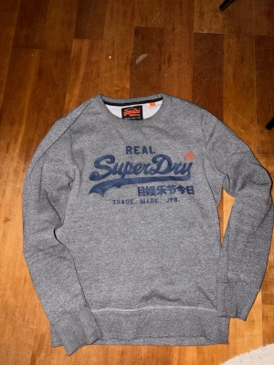 Grå sweatshirt från Superdry - Säljer en grå sweatshirt från Superdry i storlek M. Tröjan har ett stort blått tryck med Superdry-logga och japanska tecken på bröstet. Mjuk insida och ribbade muddar vid ärmslut och nederkant. Perfekt för chill dagar och streetwear.