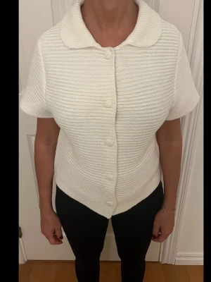 Vit stickad kofta med krage - Säljer en vit stickad kofta med korta ärmar och bred krage. Koftan har ribbad struktur och stängs med fem matchande knappar framtill. Passformen är normal och materialet känns mjukt och bekvämt. Perfekt för dig som gillar klassisk och enkel stil.
