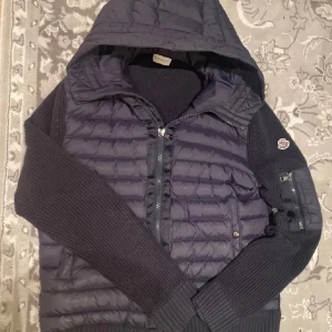 Mörkblå dunväst med huva från Moncler - Säljer en mörkblå dunväst från Moncler med stickade ärmar och huva. Västen har quiltad framsida, dragkedja och en Moncler-logga på ärmen. Materialet är en mix av dun och stickad polyester, perfekt för kyliga dagar. Snygg och sportig look med praktiska fickor.