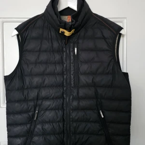 Svart quiltad dunväst med dragkedja - Säljer en svart quiltad dunväst med hög krage och snyggt spänne i halsen. Västen har tre praktiska fickor med dragkedja och diskreta orangea detaljer vid axlarna. Perfekt att slänga över en hoodie eller tröja när det är kyligt ute.