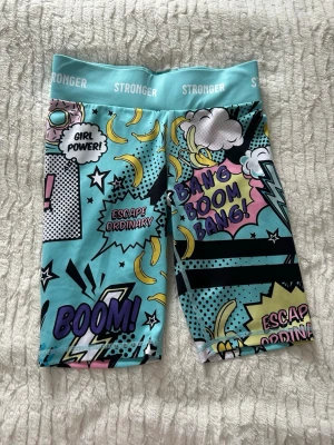 Turkosa träningsshorts från Stronger - Säljer ett par färgglada träningsshorts från Stronger i storlek S. De har ett coolt pop art-mönster med text, bananer och moln i turkos, gul, rosa, svart och vit. Hög midja och stretchigt material av återvunnen polyester och elastan. Perfekta för gymmet eller löpning!