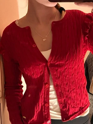 Röd kofta från Hampton Republic - Röd kabelstickad kofta från Hampton Republic med söta volangdetaljer på axlarna och röda knappar framtill. Koftan har lång ärm och en liten guldfärgad logga nertill. Perfekt för att ge din outfit lite extra färg. Funkar till alla säsonger, speciellt till jul men även sommarens kvällar.☀️🌄