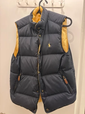 Mörkblå dunväst från Polo Ralph Lauren storlek S - Säljer en mörkblå dunväst från Polo Ralph Lauren med gul insida och klassisk logga på bröstet. Västen har hög krage, tryckknappar och två fickor med dragkedja och läderdetaljer. Perfekt för lager-på-lager under kyliga dagar.     På bild två ser man litet hål vid vänster bröst.