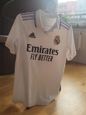 Vit Real Madrid t-shirt från Adidas - Säljer en vit Real Madrid t-shirt från Adidas med broderat klubbmärke och lila ränder på axlarna. Tröjan har korta ärmar, krage och tryck med Emirates Fly Better på bröstet. Perfekt för fotbollsfans som vill ha en snygg och sportig look. T-shirten har ett litet hål på axeln men det syns inte så mycket