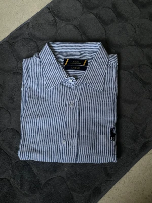 Randig skjorta från Polo Ralph Lauren - Snygg skjorta från Polo Ralph Lauren i storlek Large. Skjortan är vit med mörkblå smala ränder och har klassisk krage, knappar framtill och broderad logga på bröstet. Tillverkad i mjuk bomull som känns skön mot huden. Perfekt för dig som gillar stilrena och tidlösa plagg.