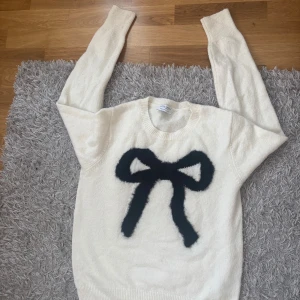 Vit stickad tröja med svart rosett - Säljer en vit stickad tröja från Holly&Whyte  med en stor svart rosett framtill. Tröjan har rund hals och långa ärmar med ribbade muddar. 