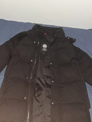 Svart dunjacka från Canada Goose - Säljer en svart dunjacka från Canada Goose med huva och klassisk logga på ärmen. Jackan har dragkedja och tryckknappar framtill, hög krage och flera praktiska fickor. Perfekt för kalla dagar och riktigt snygg streetstil.