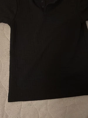 Svart slim fit topp från Black Squad - Svart topp från Black Squad i storlek L med slim fit passform. Toppen har korta ärmar och ett diskret geometriskt mönster i tyget samt knappar vid halsen. Perfekt för dig som gillar stilrena och moderna plagg.