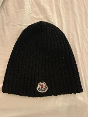 Svart Moncler mössa - Säljer en svart ribbstickad mössa från Moncler med klassisk logotyp framtill. Mössan är mjuk och skön, perfekt för kyliga dagar. Enkel och stilren design som passar till allt. Tecken på användning inget äkthetsbevis.