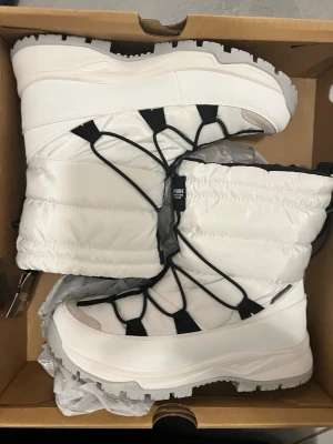 Vita vinterstövlar Icepeak strl 40 - Säljer ett par vita vinterstövlar från Icepeak, modell Ajaccio MS. Stövlarna har en quiltad, syntetisk ovandel med svarta snörningsdetaljer och grov platt sula. Insidan är svart och de har dragflik baktill. Perfekta för kalla dagar.