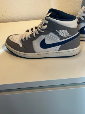 Nike Air Jordan 1 Mid grå/blå - Säljer ett par Nike Air Jordan 1 Mid i grått, vitt och marinblått. Skorna har klassisk snörning, rund tå och platt sula. Matrialet är skin och är ny tvättade och allt. Finns lite små defekter men inget som märks eller syns. Skriv vid frågor!!!