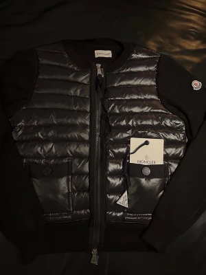 Moncler Cardigan  - Snygg svart pufferjacka från Moncler med quiltad framsida och stickade ärmar. Jackan har rund hals, två fickor med knappstängning och dragkedja framtill. Moncler-logga på ärmen och coola detaljer som ger en exklusiv vibe.