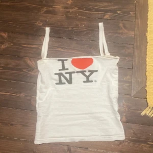 Vit I ❤️ NY linnetopp - Säljer en klassisk vit linnetopp med det ikoniska 'I ❤️ NY'-trycket framtill. Toppen har smala axelband och är perfekt för varma dagar. Enkel och stilren design som passar till mycket.