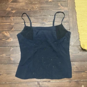 Svart linnetopp med broderi - Säljer en svart linnetopp med tunna axelband och diskret broderat mönster framtill. Toppen har smockade partier vid bysten för extra komfort och en feminin siluett. Perfekt att styla med jeans eller kjol för en trendig look.