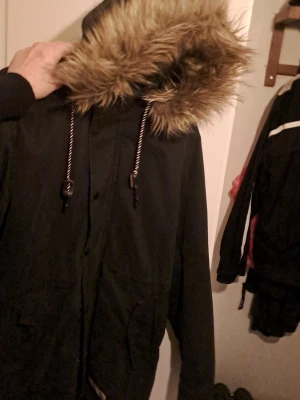 Svart parkajacka från Tommy Jeans - Säljer en svart parkajacka från Tommy Jeans med huva och dragsko. Jackan har dragkedja och knappar framtill, stora fickor och långa ärmar. Perfekt för kyligare dagar och har en klassisk, avslappnad look.