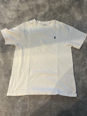 Vit t-shirt från Polo Ralph Lauren - Klassisk vit t-shirt från Polo Ralph Lauren med den ikoniska mörkblå broderade loggan på bröstet. Lite defekter som minimala hål vid midjan men man märker knappast. Pris är ej hugget i sten.