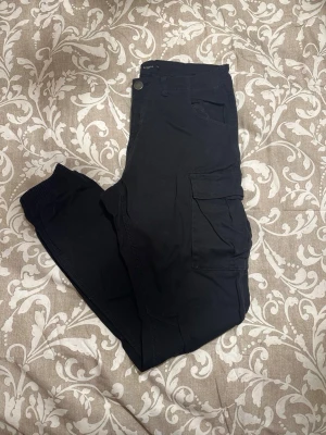 Svarta cargopants från Lager 157 - Svarta cargopants från Lager 157 i storlek S. Byxorna har flera fickor, bland annat två stora på sidorna, och resår vid bensluten för en skön passform. Materialet är mjukt och känns som bomull, perfekt för en chill och avslappnad stil.
