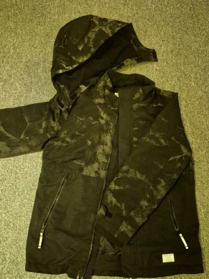 Svart vindjacka med camouflagereflex  - Svart vindjacka från H&M med coolt camouflage-mönster och huva som är reflekterande. Jackan har två dragkedjeförsedda fickor och har använts som skidjacka. Materialet känns slitstarkt och jackan har en rak passform.