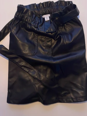 Svart kort kjol i skinnimitation Primark - Säljer en svart kort kjol från Primark i skinnimitation med hög midja och bred resår. Kjolen har stora fickor framtill och ett matchande bälte som knyts i midjan. Perfekt för dig som vill ha en edgy och trendig look.