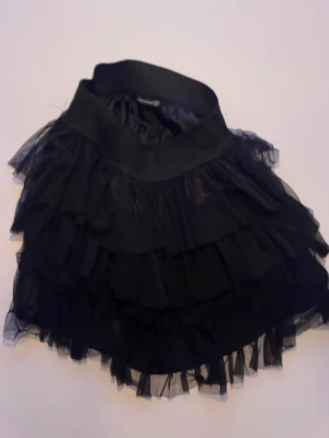 Svart volangkjol med bred midja - Säljer en svart kortkjol med flera lager volanger och bred midja. Kjolen har en fluffig och lekfull look tack vare det tunna, transparenta tyget i volangerna. Perfekt för dig som gillar att sticka ut med din stil.