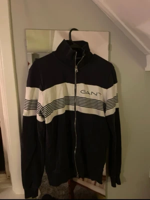 Ziptröja från GANT, S - Snygg blå och vit ziptröja från GANT i storlek S. Tröjan har hel dragkedja och ett randigt mönster över bröstet. GANT-logga framtill. Tillverkad i mjuk bomull, perfekt för chill eller när du vill ha en sportig look.